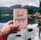 A hand holds a passport with travel-themed stamps and the words 'bon voyage' in front of a serene lake, surrounded by forested mountains under a cloudy sky.