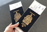 A hand holding two Canadian passports, each containing a boarding pass. The passports are navy blue with gold text and the Canadian coat of arms embossed on the cover. The background is neutral and out of focus.