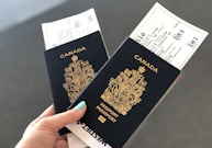 A hand holding two Canadian passports, each containing a boarding pass. The passports are navy blue with gold text and the Canadian coat of arms embossed on the cover. The background is neutral and out of focus.