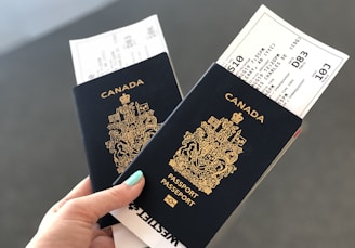 A hand holding two Canadian passports, each containing a boarding pass. The passports are navy blue with gold text and the Canadian coat of arms embossed on the cover. The background is neutral and out of focus.