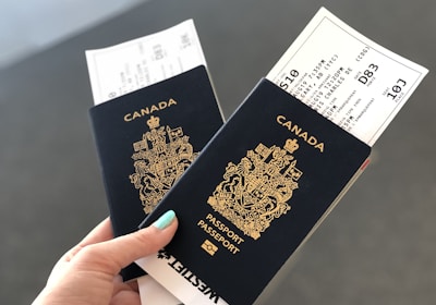 A hand holding two Canadian passports, each containing a boarding pass. The passports are navy blue with gold text and the Canadian coat of arms embossed on the cover. The background is neutral and out of focus.