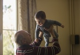 man wearing maroon, white, and blue stripe long-sleeved shirt lifting up baby wearing gray onesie