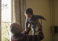 man wearing maroon, white, and blue stripe long-sleeved shirt lifting up baby wearing gray onesie