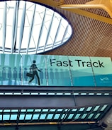An interior view of a building featuring a sign for VIP Fast Track, with a graphic of a person carrying a suitcase. The ceiling has a modern design with wooden panels and a large circular skylight.