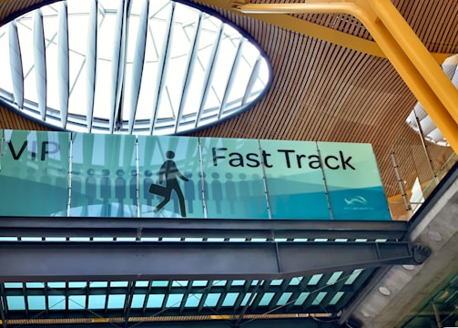 An interior view of a building featuring a sign for VIP Fast Track, with a graphic of a person carrying a suitcase. The ceiling has a modern design with wooden panels and a large circular skylight.