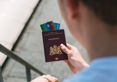 A person is holding a Dutch passport with several credit cards inserted between its pages. The passport has an emblem with a lion, and the text is written in gold. The person is wearing a light-colored shirt, and the background is slightly blurred, showing a staircase.