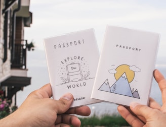 Two hands are holding up passport covers against the backdrop of a scenic lake. The left cover has an illustration of a suitcase with the words 'EXPLORE the WORLD'. The right cover features a mountain and sun illustration. There is a stone building with a rustic look on the left and flowering plants in the bottom left.
