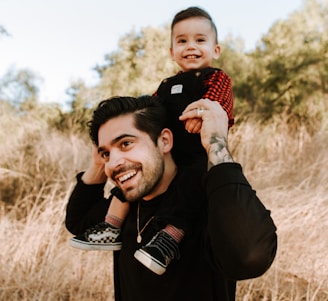 man in black jacket carrying baby in red and white checkered shirt during daytime