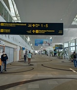 A spacious airport terminal corridor featuring a high, curved ceiling with large windows for natural light. Signs for international departures, immigration, and health services are prominently displayed. People are seen walking and pulling luggage, and a coffee shop is located to the left. The area has a modern design with patterned flooring.