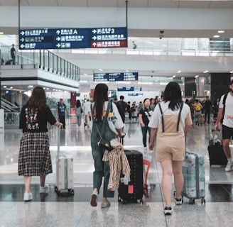 A busy airport terminal with several travelers walking and wheeling suitcases. People are moving in different directions, and overhead signs in multiple languages guide navigation. There are glass railings, escalators, and a high ceiling with bright lighting.