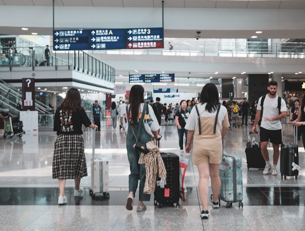 A busy airport terminal with several travelers walking and wheeling suitcases. People are moving in different directions, and overhead signs in multiple languages guide navigation. There are glass railings, escalators, and a high ceiling with bright lighting.