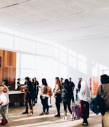 A group of people stand in line inside a bright and spacious building with large windows. The sunlight creates shadows on the floor, and some individuals carry luggage and wear backpacks. There is a counter with staff assisting the individuals. The interior features wood paneling and minimalistic decor.