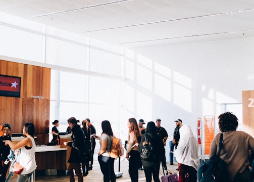 A group of people stand in line inside a bright and spacious building with large windows. The sunlight creates shadows on the floor, and some individuals carry luggage and wear backpacks. There is a counter with staff assisting the individuals. The interior features wood paneling and minimalistic decor.