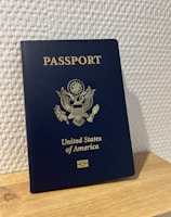 A United States passport stands upright on a wooden surface against a textured white wall. The passport is dark blue with gold embossed text and emblem on the cover.