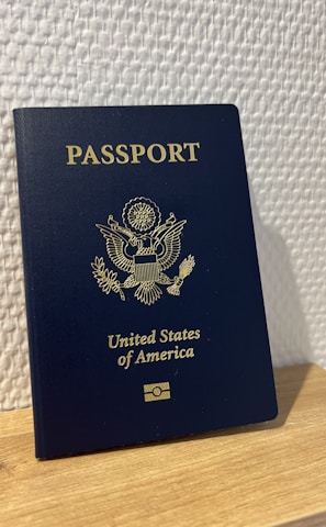 A United States passport stands upright on a wooden surface against a textured white wall. The passport is dark blue with gold embossed text and emblem on the cover.